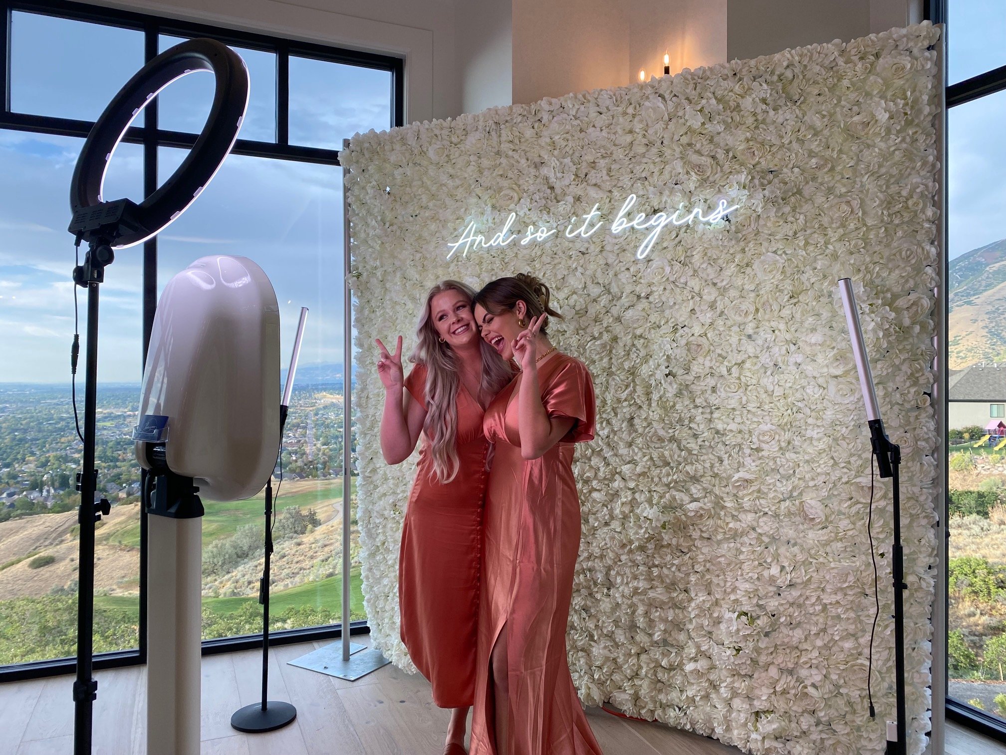 Two guests in satin dresses posing at the booth in front of the white floral wall and neon sign