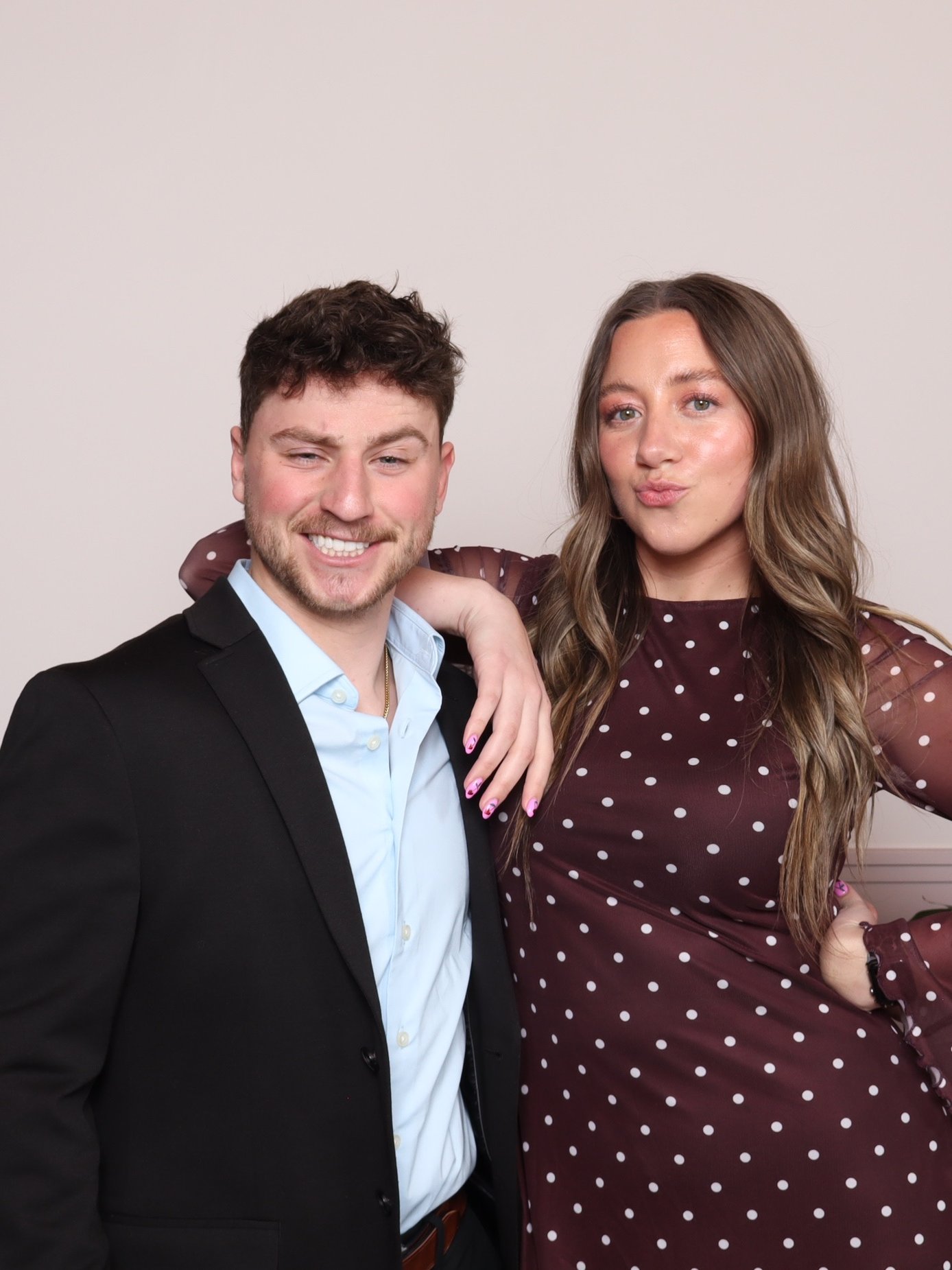 Man in a dark blazer and woman in a polka dot dress pose against a plain pale backdrop