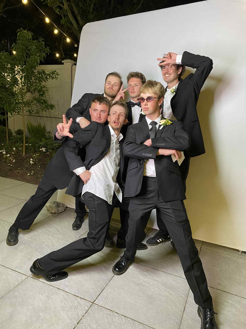 Six men in formal wear strike playful poses before a white backdrop under outdoor string lights