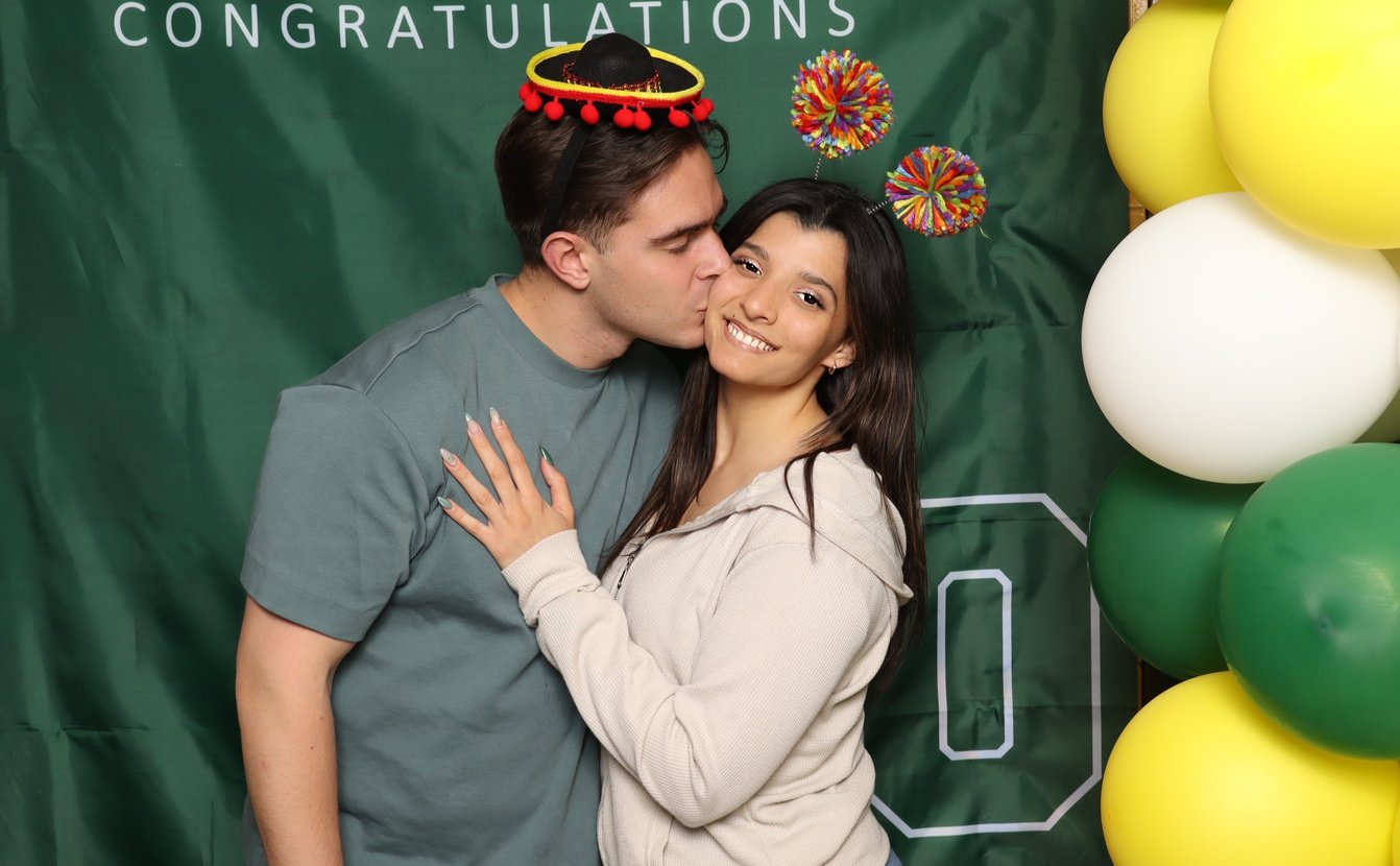 A man in a mini sombrero kisses a smiling woman's cheek before a green Congratulations backdrop and balloons
