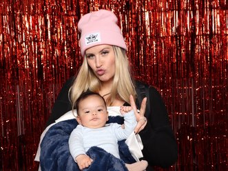 A guest holding a baby in front of a red tinsel wall at a December company party