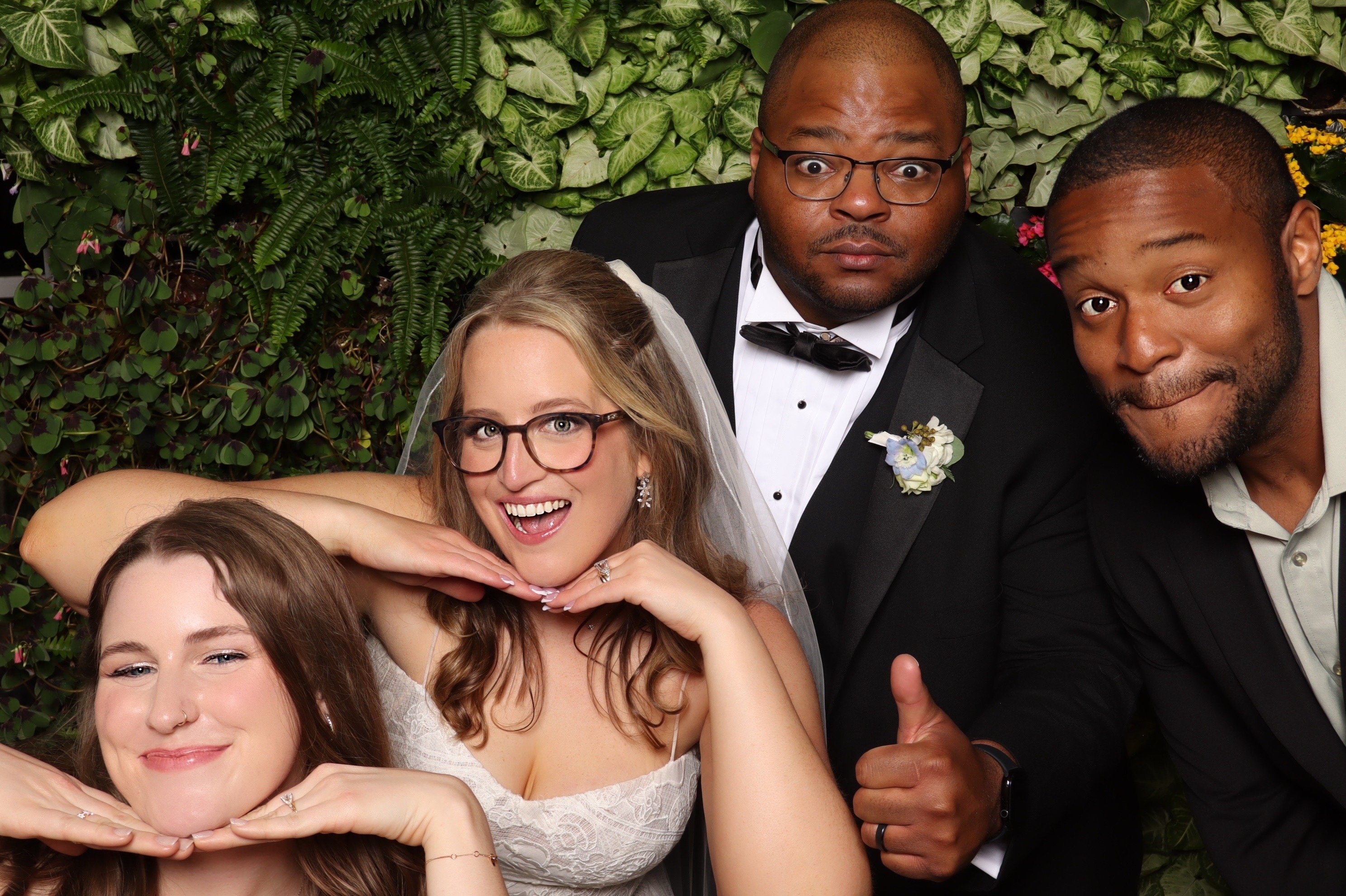 A bride and groom leaning into the frame with their guests at a greenery wall
