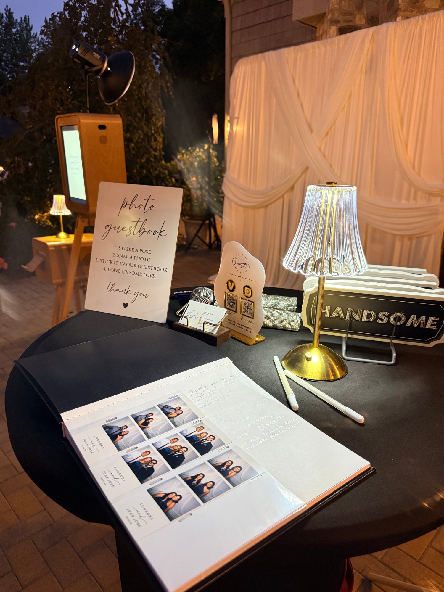 Photo guestbook table with booth strips, a sign and a gold lamp, wooden booth and white drapes behind