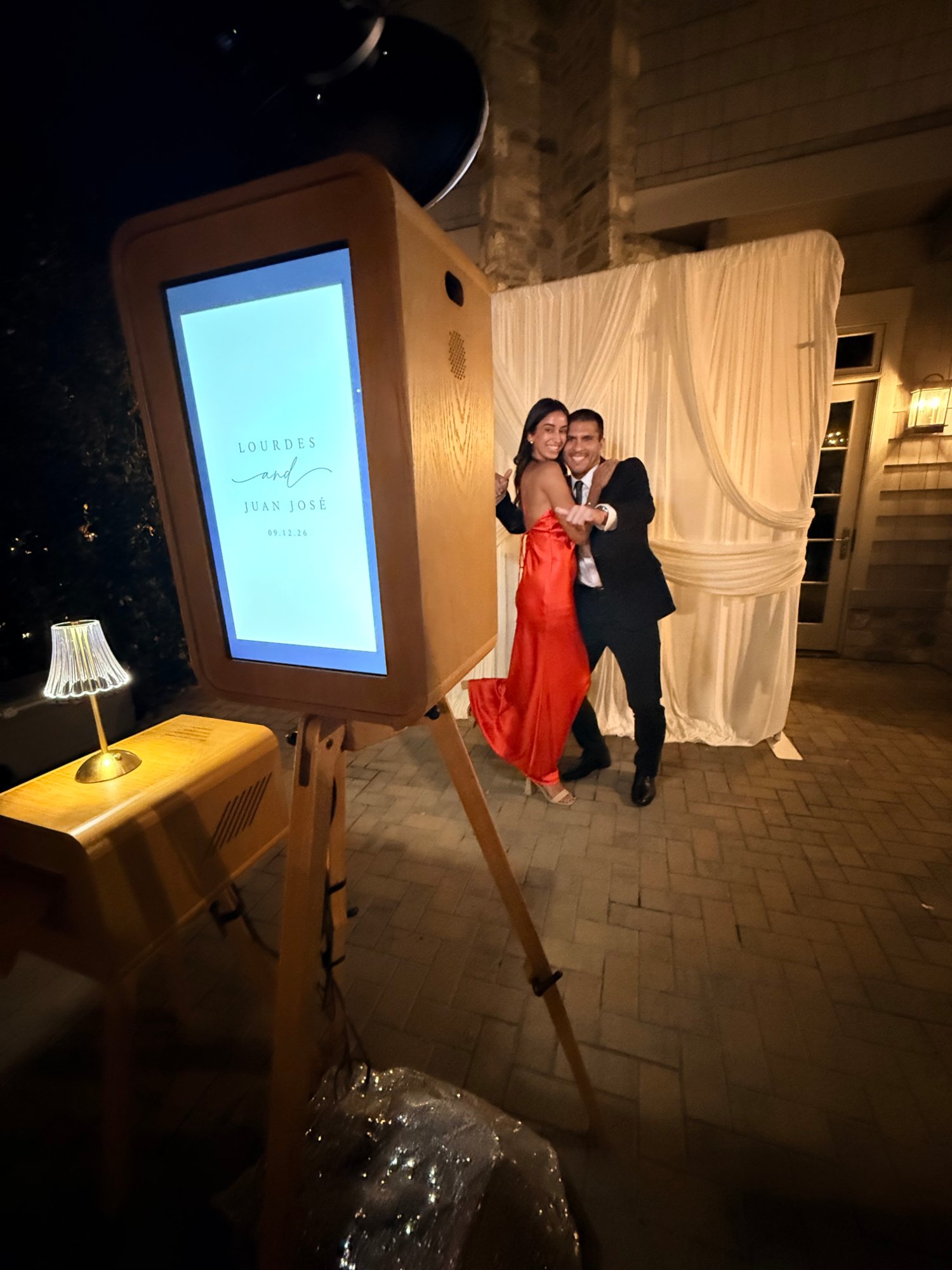 Wooden photo booth on an easel as a couple in a red gown and suit pose before white drapes